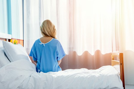 Blonde woman wearing a blue robe in hospital sitting on her bed facing the window